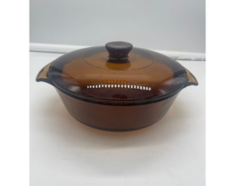 May include: A brown glass casserole dish with a lid and a wooden knob. The dish is round and has a slight lip around the edge.