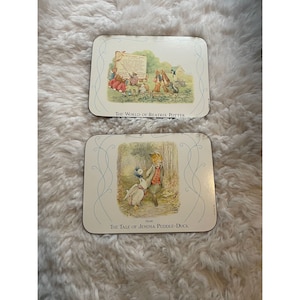 May include: Two Beatrix Potter themed coasters. The top coaster features a scene from "The Tale of Peter Rabbit" with Peter Rabbit and other rabbits. The bottom coaster features a scene from "The Tale of Jemima Puddle-Duck" with Jemima Puddle-Duck and a fox.