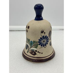 May include: A ceramic bell with a dark blue handle and a beige body. The bell is decorated with blue and green floral designs and a dark brown base. The bell is a decorative item.