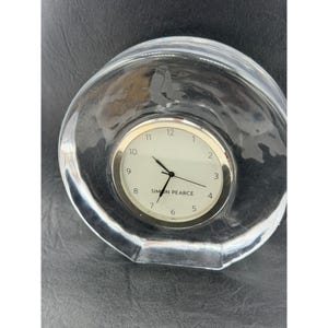 May include: A clear glass clock with a silver-colored bezel and white clock face. The clock face has black numbers and hands. The clock is encased in a clear, rounded glass structure. The brand name "SIMON PEARCE" is printed on the clock face.