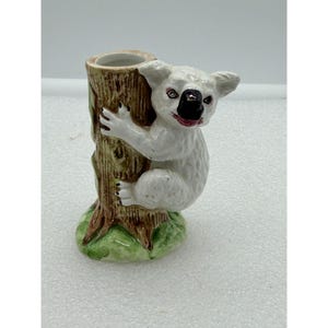 May include: A white ceramic koala bear figurine clinging to a brown tree trunk. The koala has black eyes and a black nose with a red mouth. The base is green. The figurine is a decorative item.