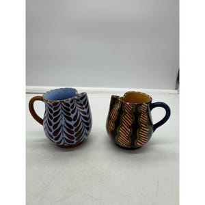 May include: Two ceramic pitchers with unique patterns. One pitcher is blue with brown and white leaf designs. The other pitcher is yellow with orange, green, and black wavy designs.