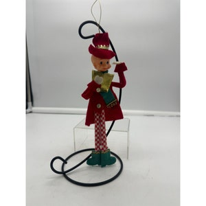 May include: A vintage Christmas ornament featuring a red-clad elf with a green scarf and a red hat with a gold crown. The elf is holding a white object in one hand and a white paper in the other. The ornament is attached to a black metal stand.