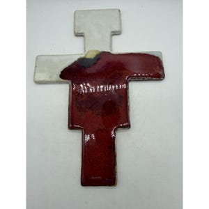 May include: A ceramic cross with a glossy finish, featuring a white upper section and a deep red lower section. The transition between the white and red glaze includes a small patch of dark brown and tan, creating a distinct color contrast.