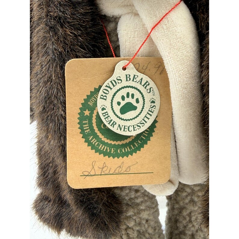 May include: A plush bear with brown and white fur, featuring a tag from "Boyds Bears" with a paw print logo. The tag reads "The Archive Collection" and has the name "Skido" written on it.
