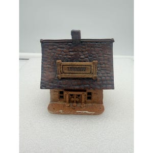 May include: A ceramic miniature library building with a brown brick-like roof and a tan facade. The word "LIBRARY" is displayed on a sign above the entrance. The building has small windows and a chimney.