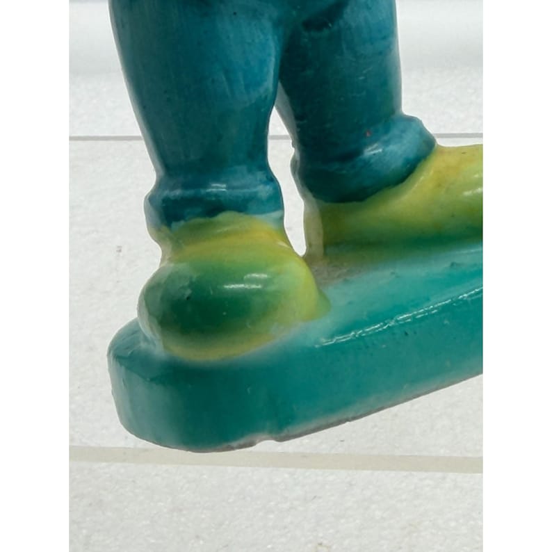 Puede incluir: Primer plano de las piernas y los pies de una figura de cer&aacute;mica. Las piernas de la figura son de color turquesa y los pies amarillos. La figura est&aacute; sobre una base turquesa. La figura est&aacute; sobre una superficie blanca.