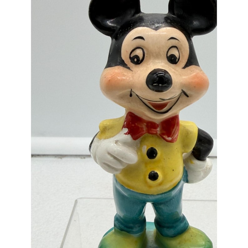 Puede incluir: Una figura vintage de Mickey Mouse. La figura tiene orejas negras, una nariz negra y una cara sonriente con mejillas rosadas. Viste una camisa amarilla, pantalones azules, una pajarita roja, guantes blancos y zapatos verdes. La figura es un objeto de colecci&oacute;n.