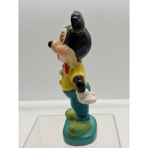 Puede incluir: Una figura vintage de Mickey Mouse. La figura tiene cabeza y orejas negras, camisa amarilla, pantalones azules y zapatos verdes. Una pajarita roja y guantes blancos completan el look. La figura se encuentra sobre una base verde azulado.