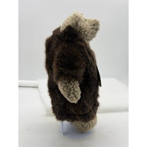 May include: A plush bear toy with a brown fur coat and tan-colored head, hands, and feet. The bear is standing on a white surface. The toy has a tag attached to it.
