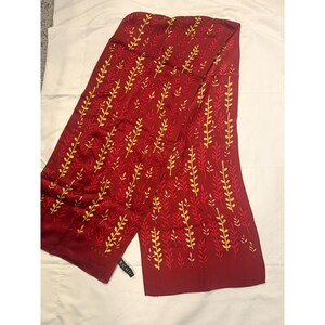 May include: A burgundy scarf with a repeating pattern of yellow and red leaf-like designs. The scarf is rectangular and appears to be made of a silky material. The brand name "ECHO" is visible on a tag at the end of the scarf.