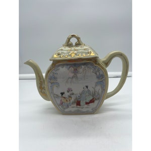 May include: A white porcelain teapot with a gold trim and a painted scene of two figures in a garden. The teapot has a square body and a lid with a floral design.