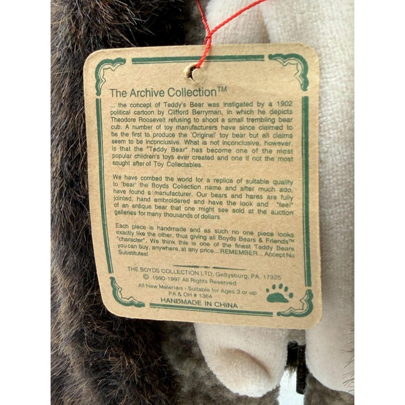 May include: A close-up of a plush teddy bear with a tag from The Archive Collection. The tag has text about the bear's history and manufacturing. The bear is handmade in China.