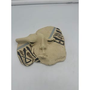 May include: A beige ceramic wall mask with a stylized face and black, brown, and gray abstract designs. The mask is signed "Lorraine" in the lower right corner.