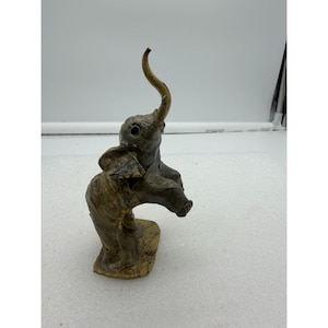 May include: A small, handcrafted elephant figurine in shades of gray and brown. The elephant has a raised trunk and large ears. The figurine is standing on a small, rectangular base. The overall style is rustic and artistic.