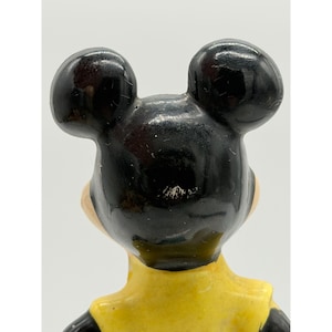 Puede incluir: Figura vintage de un personaje de dibujos animados, vista desde atr&aacute;s. La figura tiene grandes orejas negras redondas, una cara bronceada y una chaqueta amarilla. La figura probablemente est&aacute; hecha de cer&aacute;mica o un material similar.
