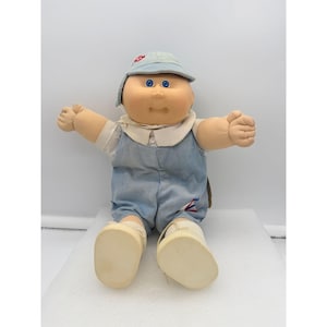 May include: A vintage Cabbage Patch Kid doll, featuring a soft, round face with blue eyes and a small nose. The doll wears a light blue corduroy overall, a white collared shirt, and a matching cap. The doll is sitting with arms outstretched.