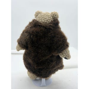 May include: A plush teddy bear with a brown fur coat and a beige head and paws. The bear is standing on a white stand, showcasing its back. The fur coat has a shaggy texture, adding to the toy's charm.