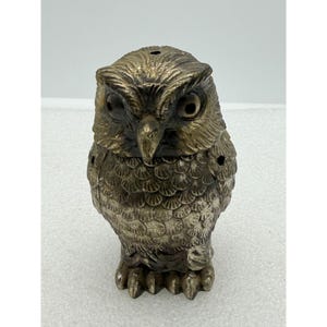 May include: A metallic owl figurine with a weathered bronze finish, featuring detailed feather textures, large circular eyes, and a sharp beak. The owl is depicted in a standing position with visible small holes on the top and sides of the body.