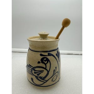 May include: A beige ceramic honey pot with a lid and a wooden honey dipper. The pot features a blue bird and floral design. The pot is approximately 5 inches tall.