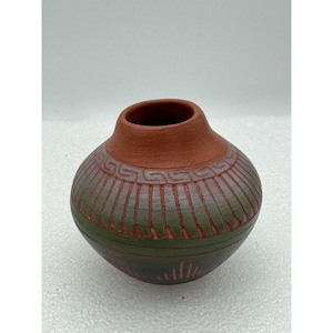 May include: A small, handcrafted terracotta pot with a rounded body and a narrow opening. The pot features a reddish-brown rim and a green-toned body with etched geometric patterns. The base has a series of vertical lines radiating outwards.