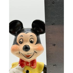 Puede incluir: Una figura vintage de Mickey Mouse. La figura tiene cabeza y orejas negras, una camisa amarilla y una pajarita roja. La figura mide aproximadamente 13 cm de alto, seg&uacute;n la regla de la imagen.
