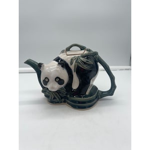 May include: A ceramic teapot shaped like a panda bear sitting in bamboo. The teapot is white with black markings and has green bamboo details. The teapot has a spout and a handle.