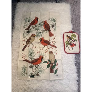 May include: A cream-colored kitchen towel and matching pot holder featuring illustrations of red cardinals perched on branches with pine needles and berries. The pot holder has a red border.