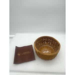 Puede incluir: Una pequeña cesta redonda tejida con un acabado marrón claro. La cesta tiene un diseño tejido y una forma ligeramente ovalada. Un papel marrón con texto está al lado de la cesta. La cesta está vacía.