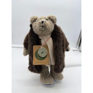 May include: A plush teddy bear wearing a brown fur coat and a cream-colored scarf. The bear has a tag that reads "Boyds Bears Archive Necessities". The bear is standing on a clear plastic base.