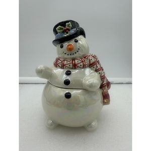 May include: A ceramic snowman figurine with a black top hat, orange nose, and a patterned scarf. The snowman has three black button details and a pearlescent finish. The figurine is a festive holiday decoration.
