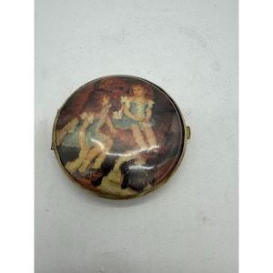 May include: A round, antique trinket box with a detailed illustration on the lid. The image depicts two young girls in blue dresses, set against a backdrop of warm, earthy tones. The box has a gold-colored metal frame.