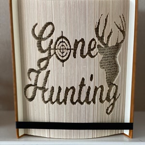 May include: A book art sculpture with the words "Gone Hunting" cut out of the pages. The sculpture features a deer silhouette and a target symbol.