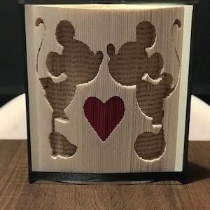 Könnte beinhalten: Ein handgefertigtes Buchkunstwerk mit Silhouetten von zwei Zeichentrickfiguren, die sich gegenüberstehen, und einem roten Herz in der Mitte. Die Seiten des Buches sind gefaltet und geschnitten, um das Design zu erstellen, in einem schwarzen Rahmen.