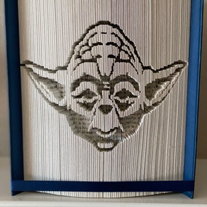 以下が含まれることがあります： スターウォーズのヨーダのブックアート彫刻。彫刻は、ページを折り畳んで画像を作成した本で作られています。彫刻は青い枠と青いリボンで囲まれています。