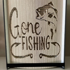 Könnte beinhalten: Eine Buchkunstskulptur mit einem Fisch und den Worten "Gone Fishing", die aus den Seiten ausgeschnitten sind. Das Buch ist in Schwarz gebunden und hat einen schwarzen Ständer.