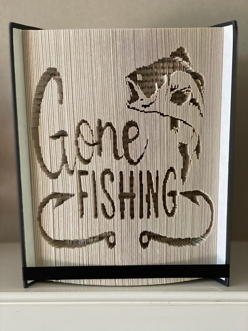 May include: A book art sculpture with a fish and the words "Gone Fishing" cut out of the pages. The book is bound in black and has a black stand.