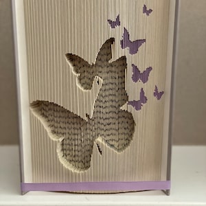 Peut inclure: Une sculpture d'art de livre représentant deux papillons découpés dans les pages d'un livre. Les papillons sont en vol et sont entourés de papillons plus petits. La sculpture est faite d'un livre avec un ruban violet autour du bas.