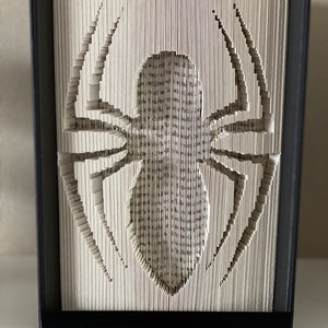 Könnte beinhalten: Eine Buchkunstskulptur einer Spinne, hergestellt durch Falten der Seiten eines Buches. Die Spinne ist aus den Seiten ausgeschnitten, wodurch ein 3D-Effekt entsteht. Die Skulptur ist in einem schwarzen Rahmen gerahmt.