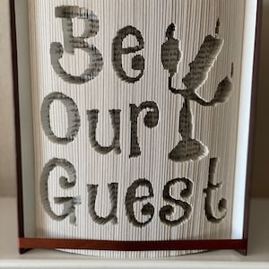 Może przedstawiać: Rzeźba książkowa z wyciętymi słowami "Be Our Guest" z kartek. Rzeźba wykonana jest z książki w brązowej okładce i brązową wstążką jako zakładką. Rzeźba stoi na białym tle.