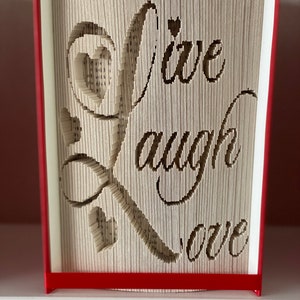 Op de afbeelding: Een boekkunst sculptuur met de woorden "Live Laugh Love" uitgesneden uit de pagina's. Het boek is gebonden in rood en de woorden zijn in een cursief schrift.