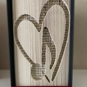 May include: A book art sculpture featuring a heart shape created by folding the pages of a book. The book is bound in black with a red ribbon at the bottom.