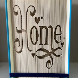 Può includere: Un libro con la parola "Home" tagliata dalle pagine, con una copertina blu e un nastro blu. Il libro è piegato in una forma tridimensionale.