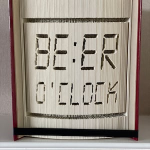 Op de afbeelding: Een boek met pagina's die zijn gevouwen in de vorm van een digitale klok die "BEER O'CLOCK" weergeeft. Het boek is gebonden in rood en heeft een zwart lint als bladwijzer.