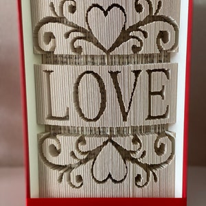 May include: A book art sculpture with the word "LOVE" in a decorative font, surrounded by intricate heart and floral designs. The book is bound in red and is standing on a white surface.