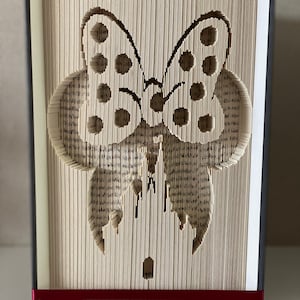 May include: A book art sculpture of a butterfly with black and white polka dots. The butterfly is made from the pages of a book that have been folded and cut to create the design. The sculpture is standing on a white surface with a red ribbon around the base.