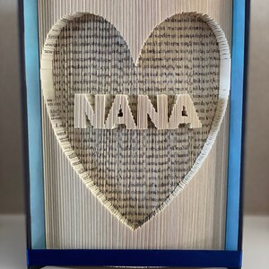 May include: A book art sculpture with a heart shape cut out of the pages. The word "NANA" is cut out of the pages in the center of the heart. The book is bound in blue fabric with a blue ribbon at the bottom.