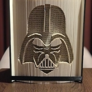 May include: A book folded into the shape of Darth Vader's head. The book is made of white paper and has black edges. The book is standing on a wooden surface.
