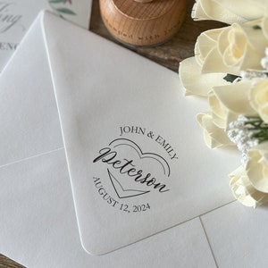 May include: A white envelope with a black and white custom wedding stamp. The stamp features a heart with the text "Peterson" and the date "August 12, 2024".