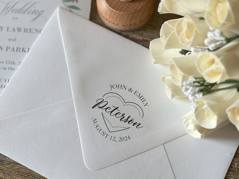 May include: A white envelope with a black and white custom wedding stamp. The stamp features a heart with the text "Peterson" and the date "August 12, 2024".
